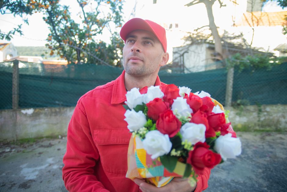 A man dressed in a red jacket and matching red cap holding a vibrant floral bouquet in front of him. The bouquet contains red and white roses with lush green leaves, arranged in a colorful patterned p