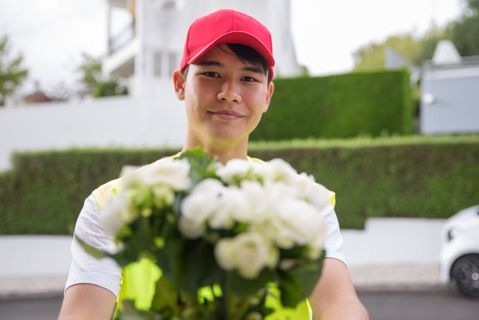 A delivery person wearing a red cap and high-visibility yellow vest extends a bouquet of fresh white flowers towards the camera, smiling gently. The bouquet is arranged with white roses and green foli