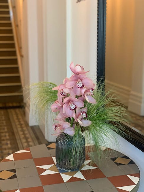 A floral arrangement featuring a tall, elegant bouquet of pale pink orchids with large, open petals and intricate white and purple markings in the centers. The orchids are set in a dark, ribbed glass 