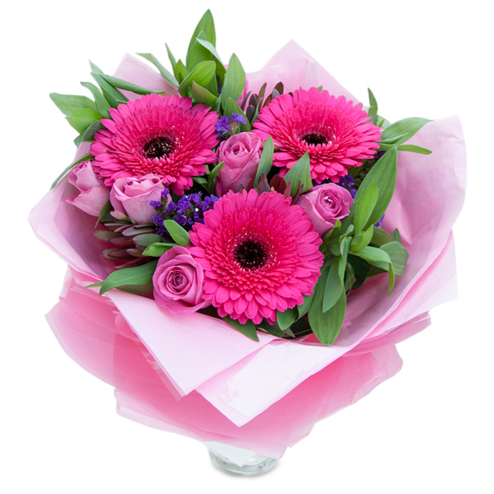 The charm of this bouquet is immediate: three confident fuchsia Gerbera daisies dominate the scene, their vivid petals spreading like cheerful suns and drawing attention to a deep, almost inky central disk. Around them, roses in a muted lavender-pink or dusty-rose hue sit as softer companions, each bud layered with velvety petals that suggest a gentle unfolding soon to come. Between these focal flowers, sprigs of purple statice peek through-tiny, papery blooms that add a textured, meadow-like sparkle and a cooler note to the warm pinks. A generous sweep of greenery-broad, dewy leaves reminiscent of salal or eucalyptus-frames the bouquet, lending structure and a fresh, slightly resinous scent. The arrangement is wrapped for gifting with care: the stems are cushioned in pale pink matte tissue, while a second, sheer layer of crinkled fuchsia-pink cellophane lends a soft sheen and completes the pink-dominant palette. The lower edge of the composition exposes the top rim of a clear glass vase, a subtle cue that the bouquet is intended for immediate display on a bedside table, hallway console, or as a welcoming centrepiece. In imagining the scene, I see the soft light of an early afternoon in Stepney casting gentle highlights across the petals, emphasising textures and the florist's attention to balance. This bouquet's scent is delicate-roses offering a faint perfume, gerberas a whisper of green-making it a tender, uplifting choice for birthdays, anniversaries, or as a thoughtful 'just because' surprise for someone special in the local community.